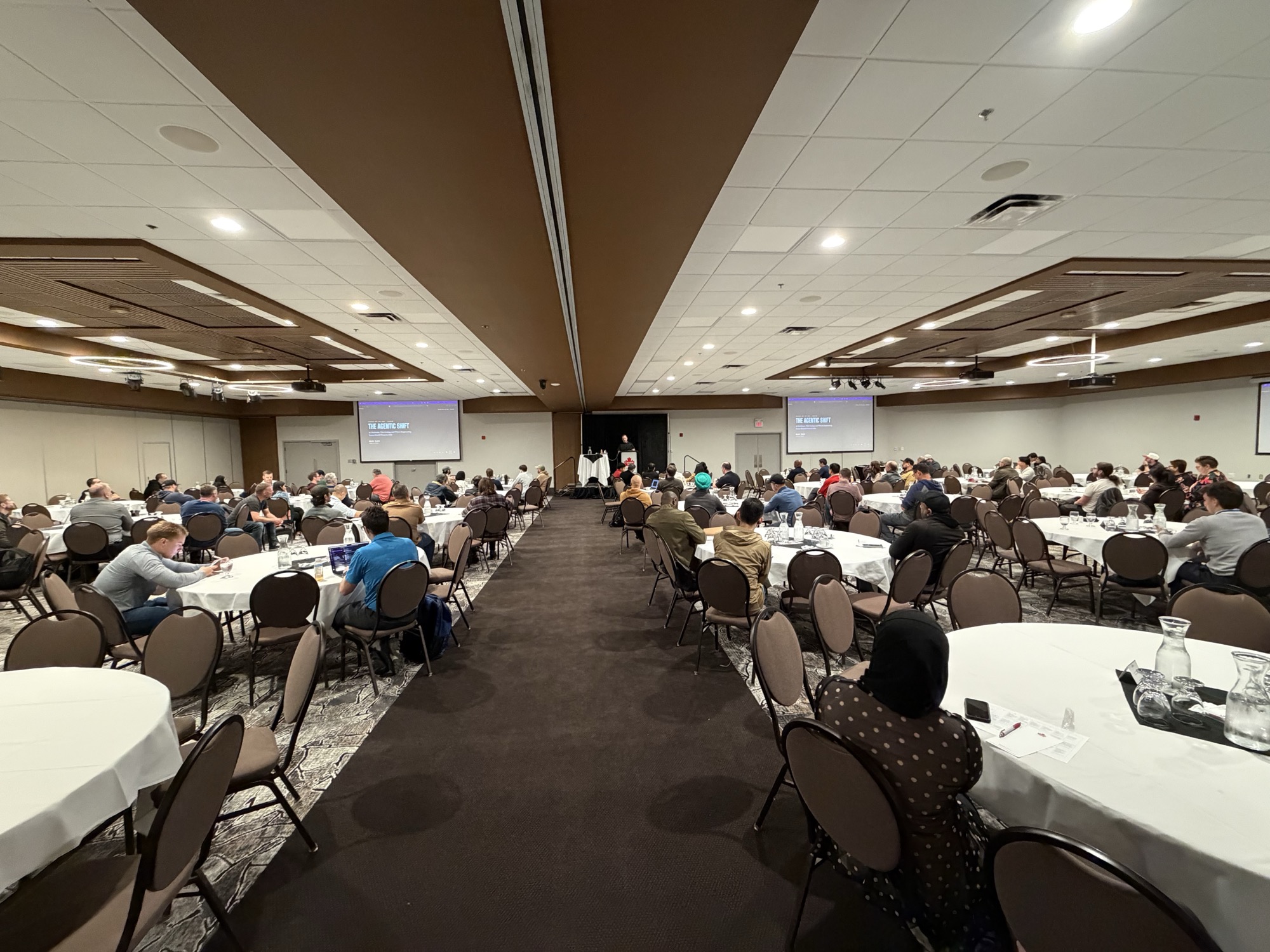 A packed conference ballroom at Prairie Dev Con Winnipeg 2026, with 'The Agentic Shift' title slide displayed on twin projector screens as Kevin Evans presents from the podium.