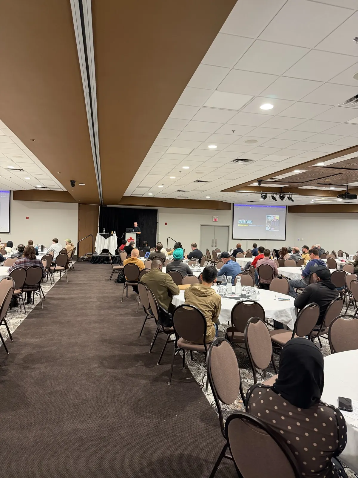 Kevin Evans presenting at a podium at Prairie Dev Con Winnipeg 2026, with a 'Who's Talking: Kevin Evans' introduction slide displayed on screen behind him.